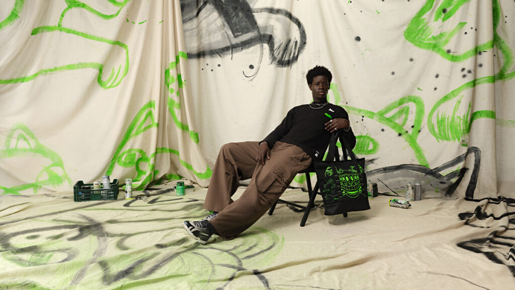 Modelo sentado em uma cadeira, usando roupas oversized e segurando uma bolsa preta, com um fundo de tecido que apresenta grafismos em verde e preto.
