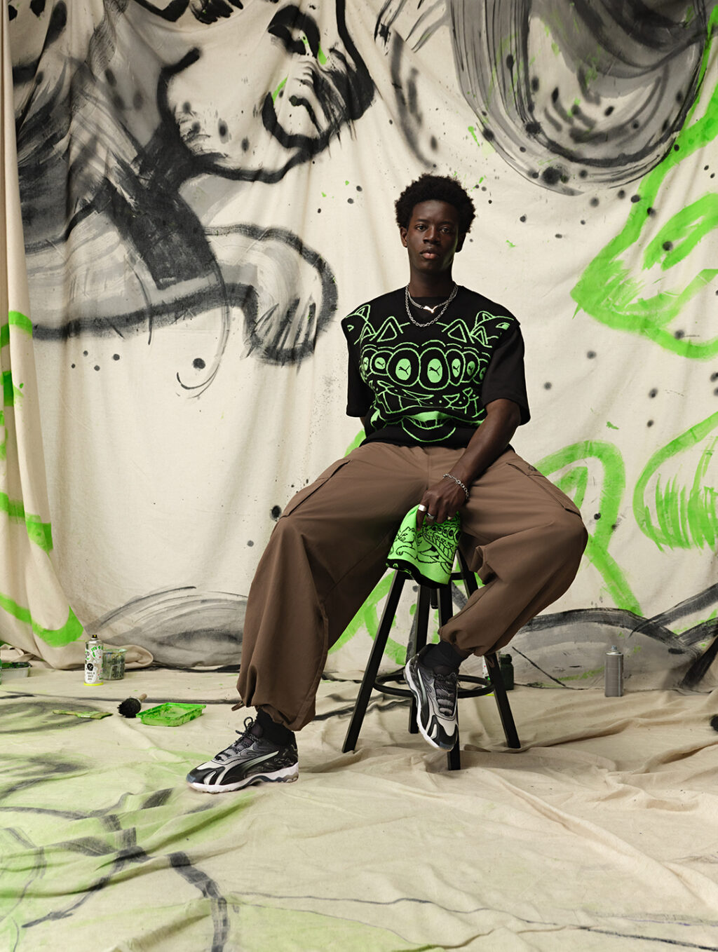 Modelo sentado em um banquinho, vestindo camiseta preta com estampa verde de gato, calças largas marrons e tênis escuros, em frente a um fundo ilustrado com grafite e tinta verde.