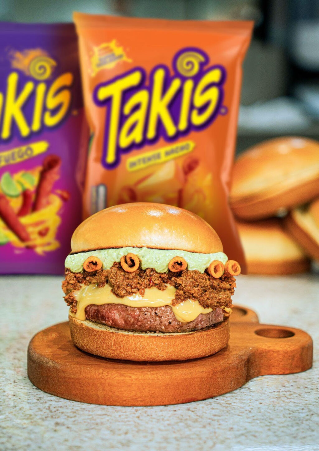 Um hambúrguer artesanal com camadas de carne, queijo e um toque crocante de Takis, posicionado em uma tábua de madeira, com pacotes de snacks Takis ao fundo.