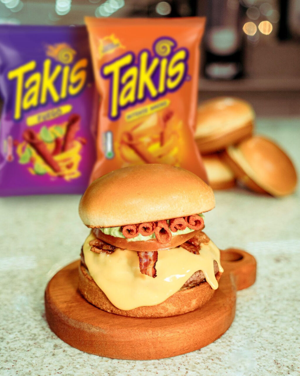 Um hambúrguer com queijo americano derretido, pedacinhos de bacon e Takis, servido em um prato de madeira, com pacotes de snacks Takis ao fundo.