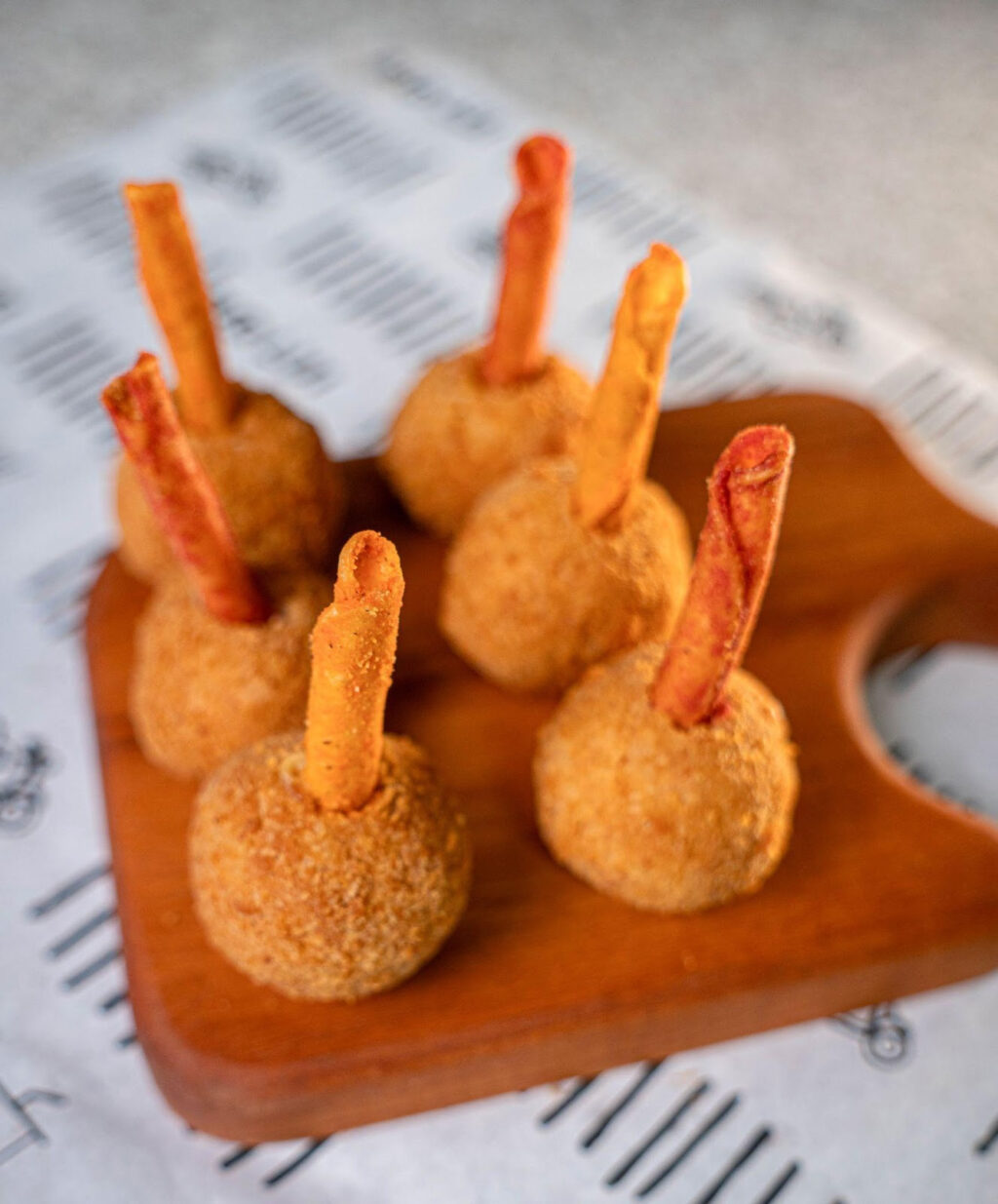Uma tábua de madeira com bolinhos empanados recheados e palitos de Takis como enfeite, sobre um fundo decorativo.