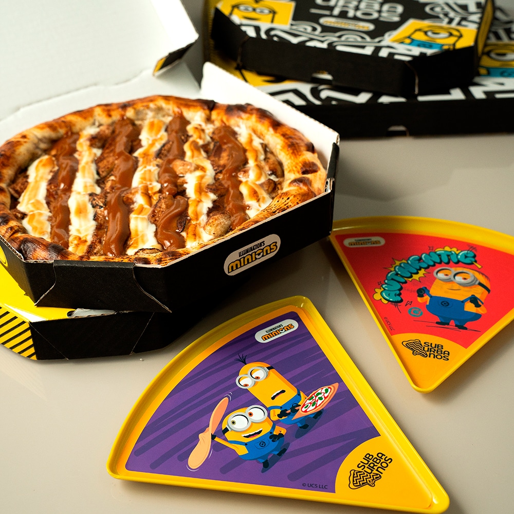 Pizza doce de banana com chocolate branco e doce de leite, acompanhada de pratos colecionáveis dos Minions em cores vibrantes.