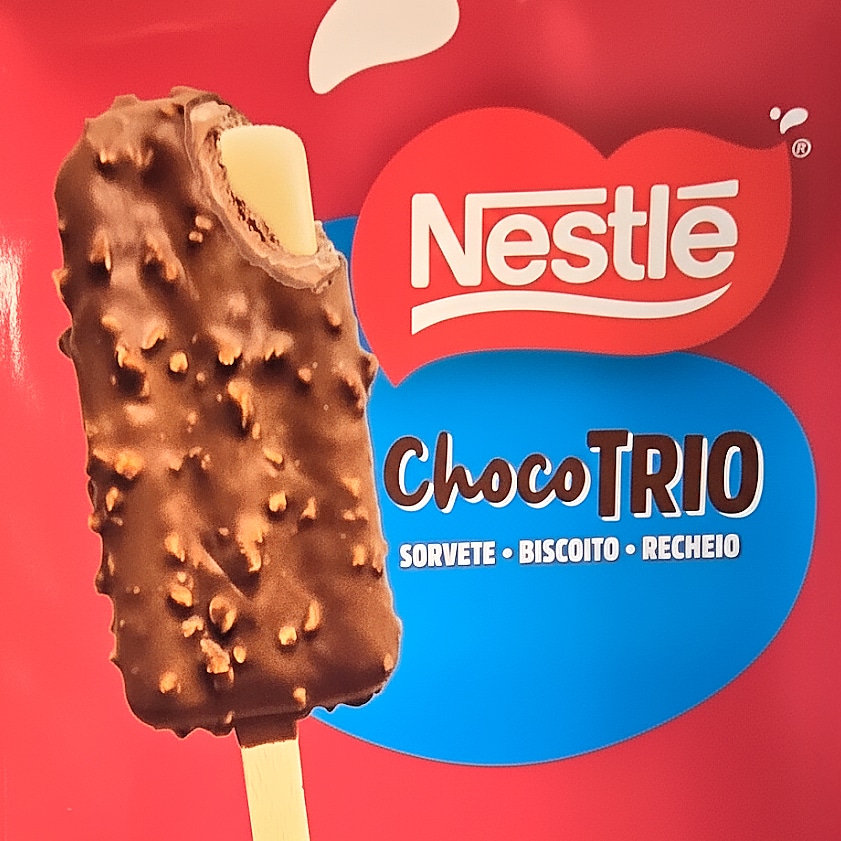 Picolé Choco Trio da Nestlé, com cobertura de chocolate e pedacinhos de biscoito, exibido em um fundo vermelho com informações sobre os sabores.