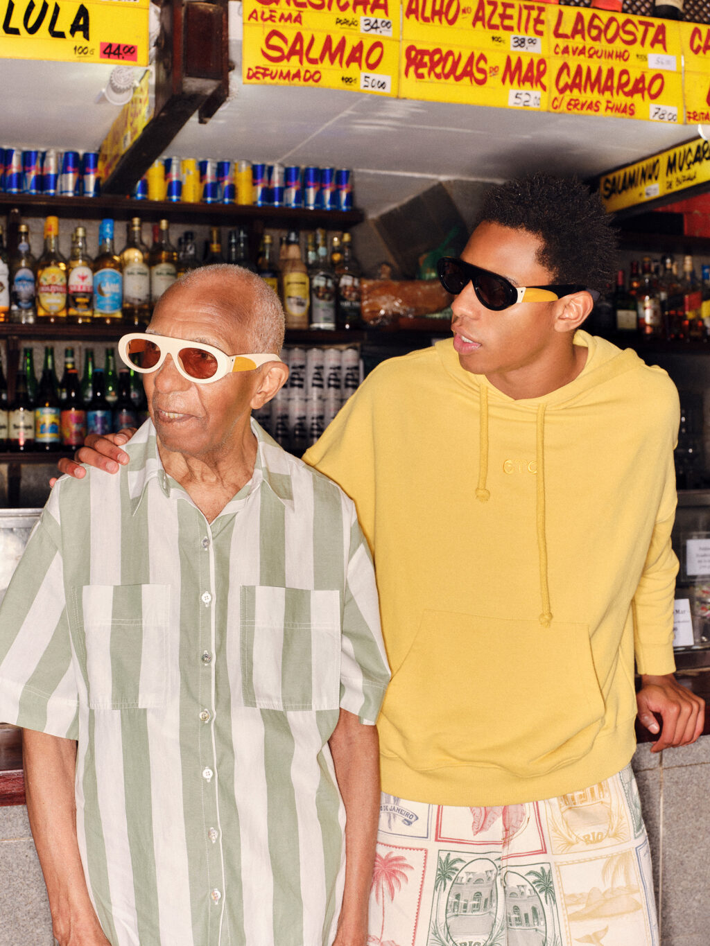 Uma foto de um avô e seu neto em um ambiente de bar, ambos vestindo óculos de sol da colaboração FARM Rio e Zerezes. O avô usa uma camisa listrada azul e verde, enquanto o neto está vestido com um moletom amarelo. Ao fundo, prateleiras com bebidas e placas de menu exibindo preços.