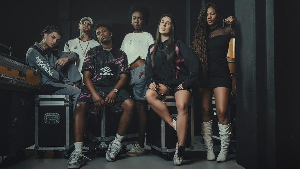 Grupo de seis pessoas posando em um ambiente interno, vestindo roupas da colaboração Umbro x KondZilla, que incluem jerseys, hoodies e acessórios. O cenário transmite uma estética urbana e de streetwear, com elementos de cultura funk.