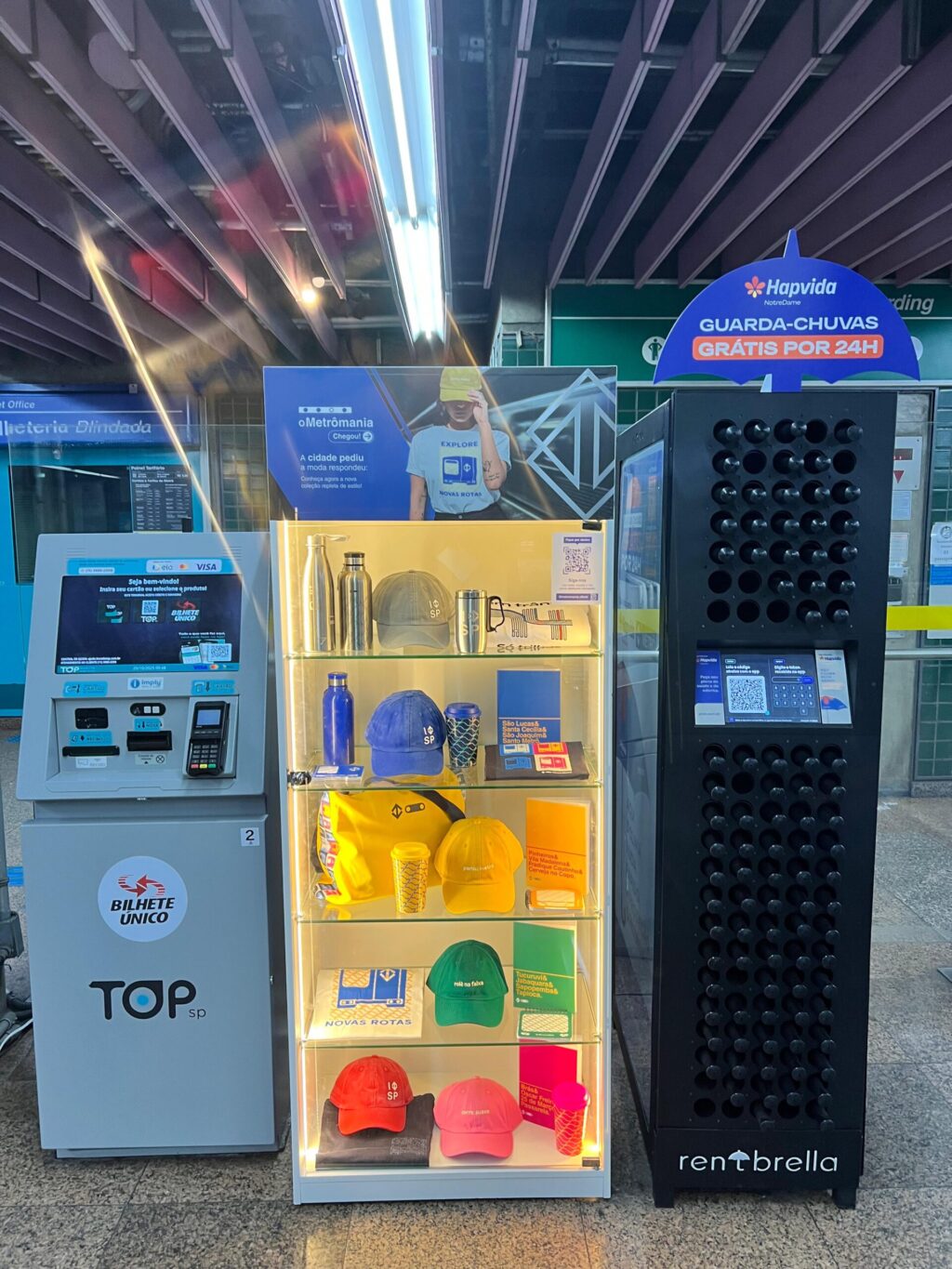Imagem do interior da estação de metrô com um quiosque da loja virtual Metrômania, mostrando produtos como garrafas, bonés e acessórios, além de um guichê de venda de bilhetes e um ponto de aluguel de guarda-chuvas.