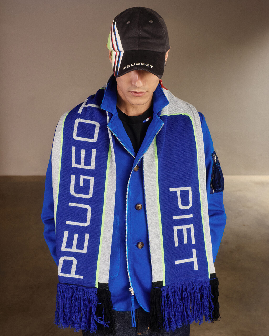 Modelo vestindo uma jaqueta azul e um cachecol com as marcas Peugeot e Piet, em um ambiente interno neutro.