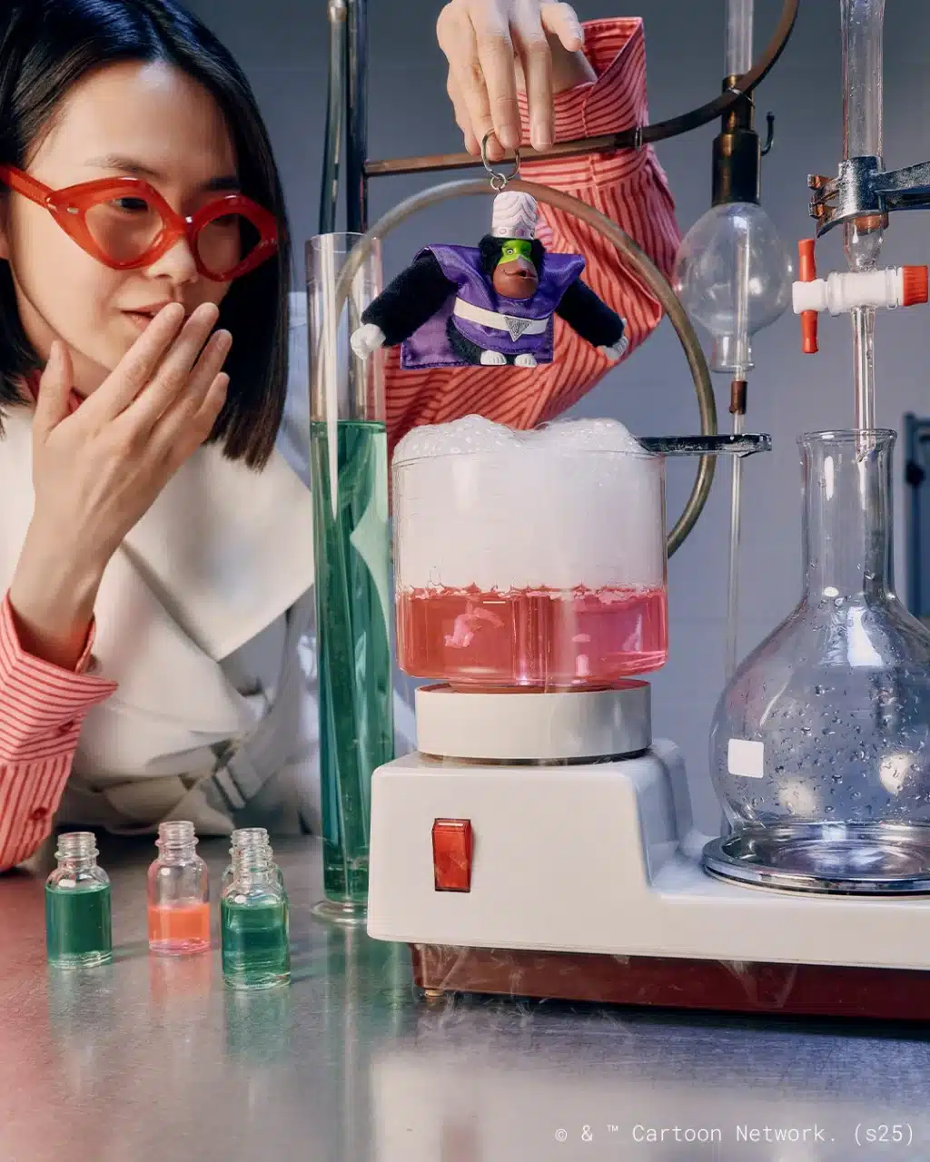 Uma mulher segurando um chaveiro do vilão Macaco Louco em um laboratório, com tubos de ensaio coloridos e um recipiente com líquido borbulhante.