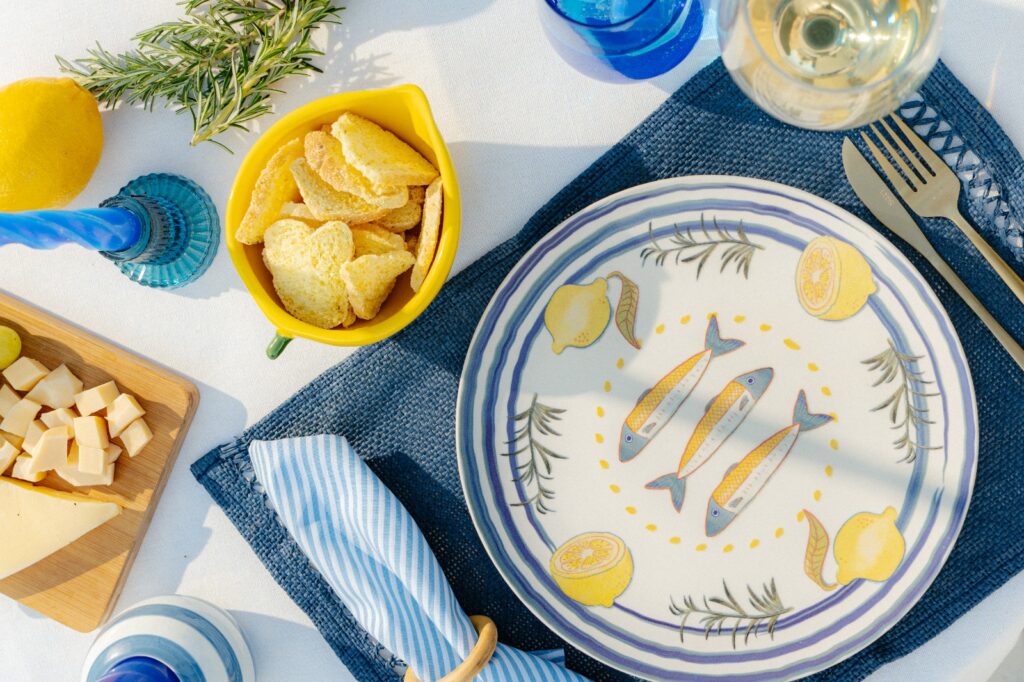 Um prato decorativo com ilustrações de peixes e limões, colocado sobre uma toalha de mesa azul. Ao lado, há uma tigela amarela com biscoitos e um tabuleiro com cubos de queijo, além de um copo de vinho e uma vela azul.