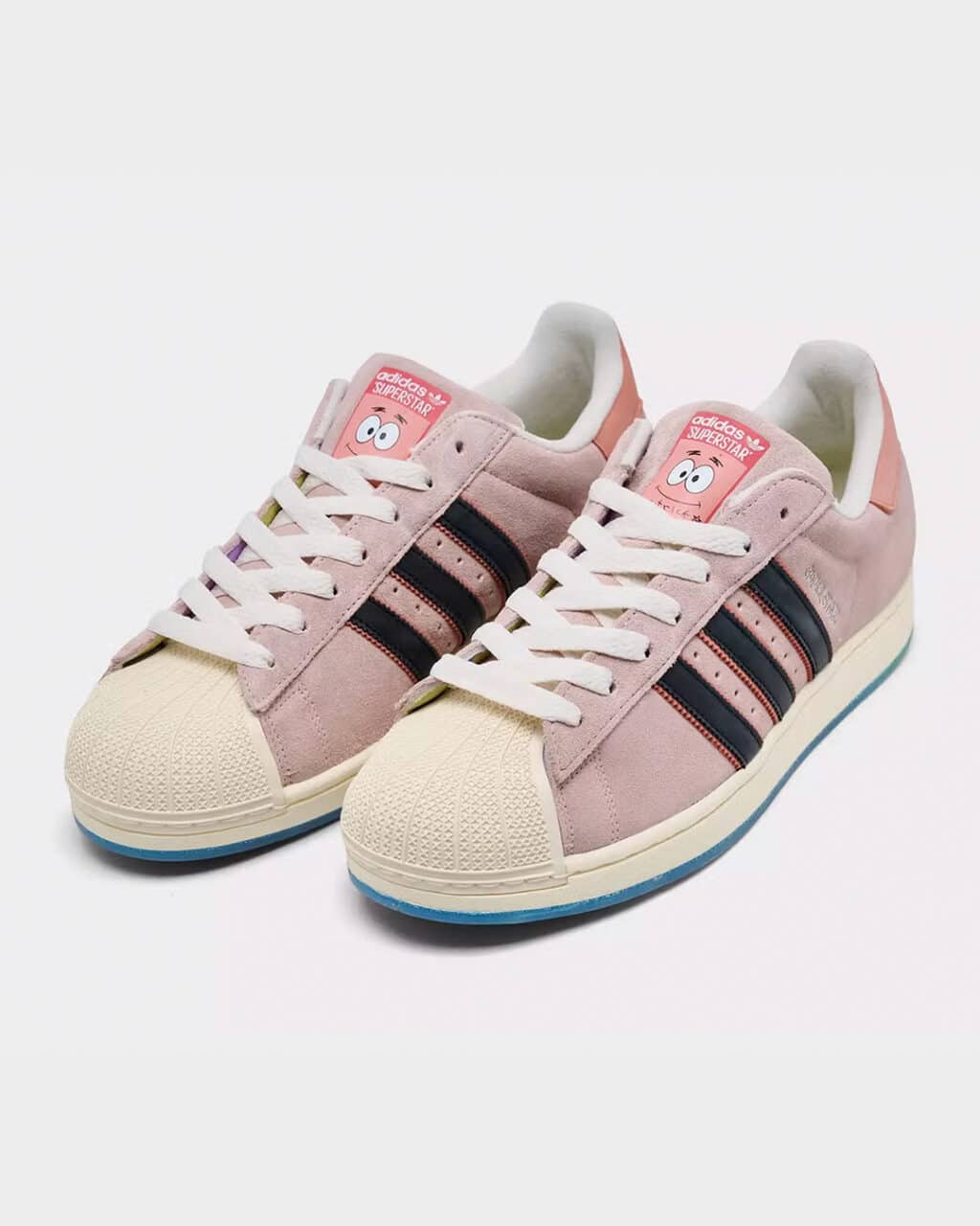 Tênis Adidas Superstar na cor rosa pastel, com cabedal em camurça, três listras pretas laterais e biqueira em borracha off-white. A língua exibe o rosto de Patrick Estrela.