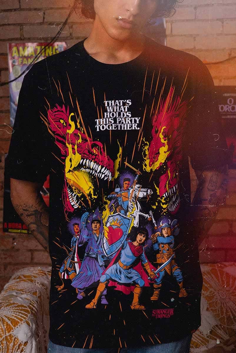Modelo vestindo uma camiseta temática de Stranger Things com ilustrações de personagens como se fossem de RPG, com a frase 'THAT'S WHAT HOLDS THIS PARTY TOGETHER'.