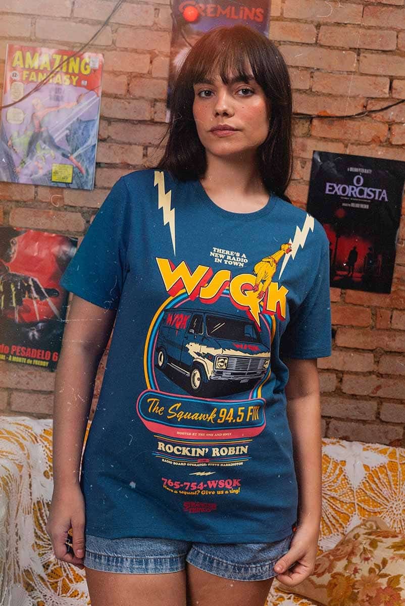 Modelo usando uma camiseta azul com a estampa da rádio WSQK de Stranger Things, em frente a uma parede de tijolos expostos e pôsteres de filmes clássicos.