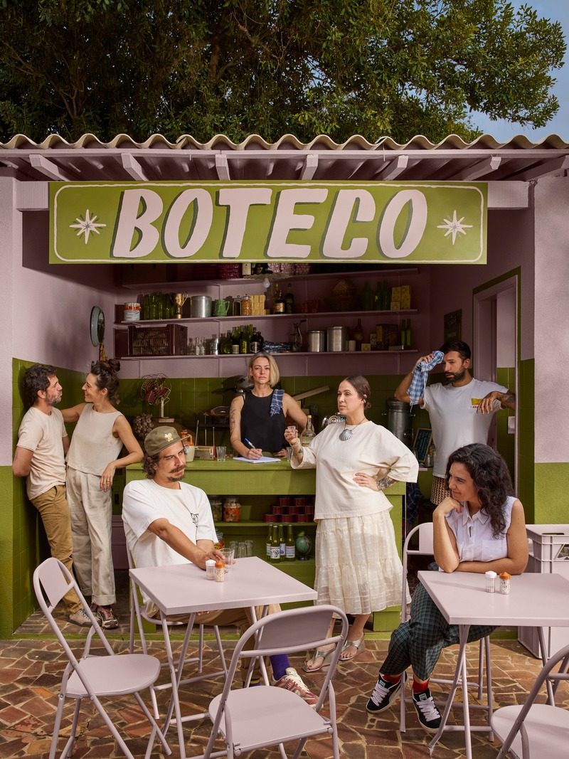 Um grupo de pessoas interagindo em um boteco com uma decoração colorida. Ao fundo, uma placa grande escrita 'BOTECO'. Algumas pessoas estão sentadas nas mesas, enquanto outras estão em pé, conversando e se divertindo.
