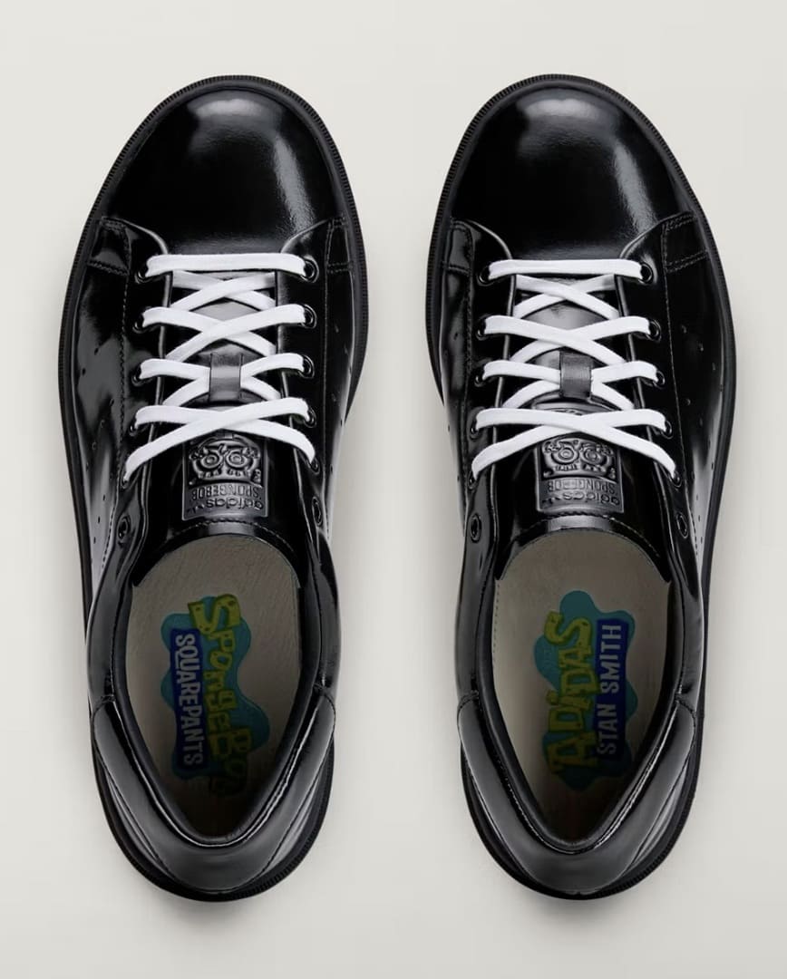 Vista superior de um par de tênis Adidas Stan Smith em couro preto brilhante com cadarços brancos, apresentando detalhes do Bob Esponja na língua e palmilhas.