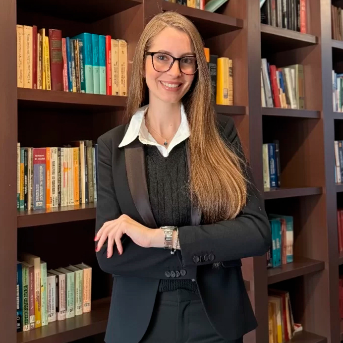 Profissional em traje formal sorrindo e com os braços cruzados, posicionada em frente a uma estante de livros.