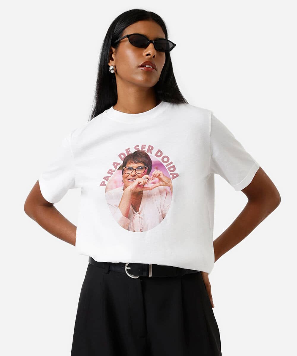 Modelo vestindo uma camiseta branca com a frase 'PARA DE SER DOIDA' e imagem de uma mulher sorridente fazendo um coração com as mãos.