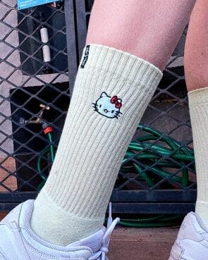 Meia de social em tom claro com bordado da personagem Hello Kitty, usada em uma perna, em um ambiente casual.
