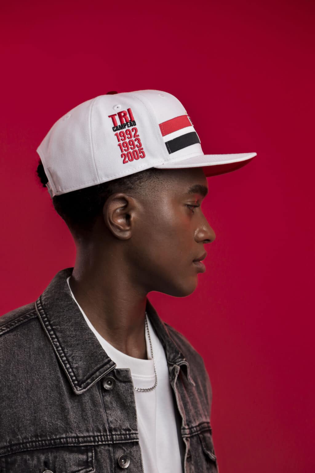 Modelo usando um boné branco com o texto 'TRI CAMPEÃO' e datas de títulos do São Paulo FC, posando contra um fundo rosa.