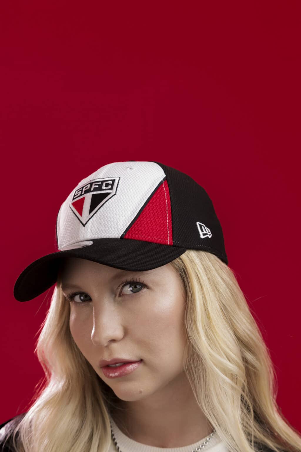 Mulher usando um boné da New Era com o logo do São Paulo FC, em frente a um fundo vermelho.