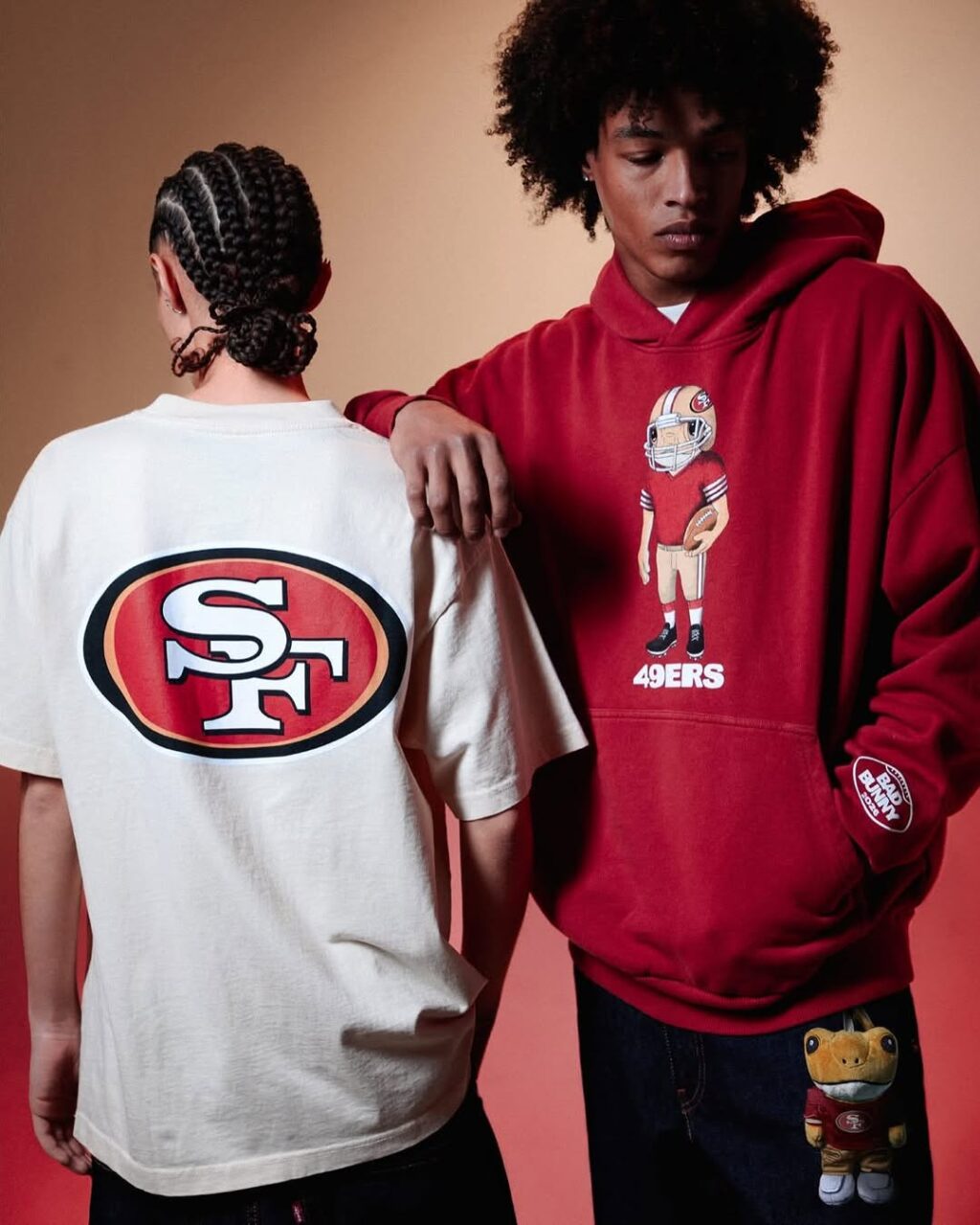 Duas pessoas usando roupas da NFL: uma com uma camiseta clara com o logotipo do San Francisco 49ers, e a outra com um moletom vermelho apresentando uma ilustração de jogador e acessórios relacionados.