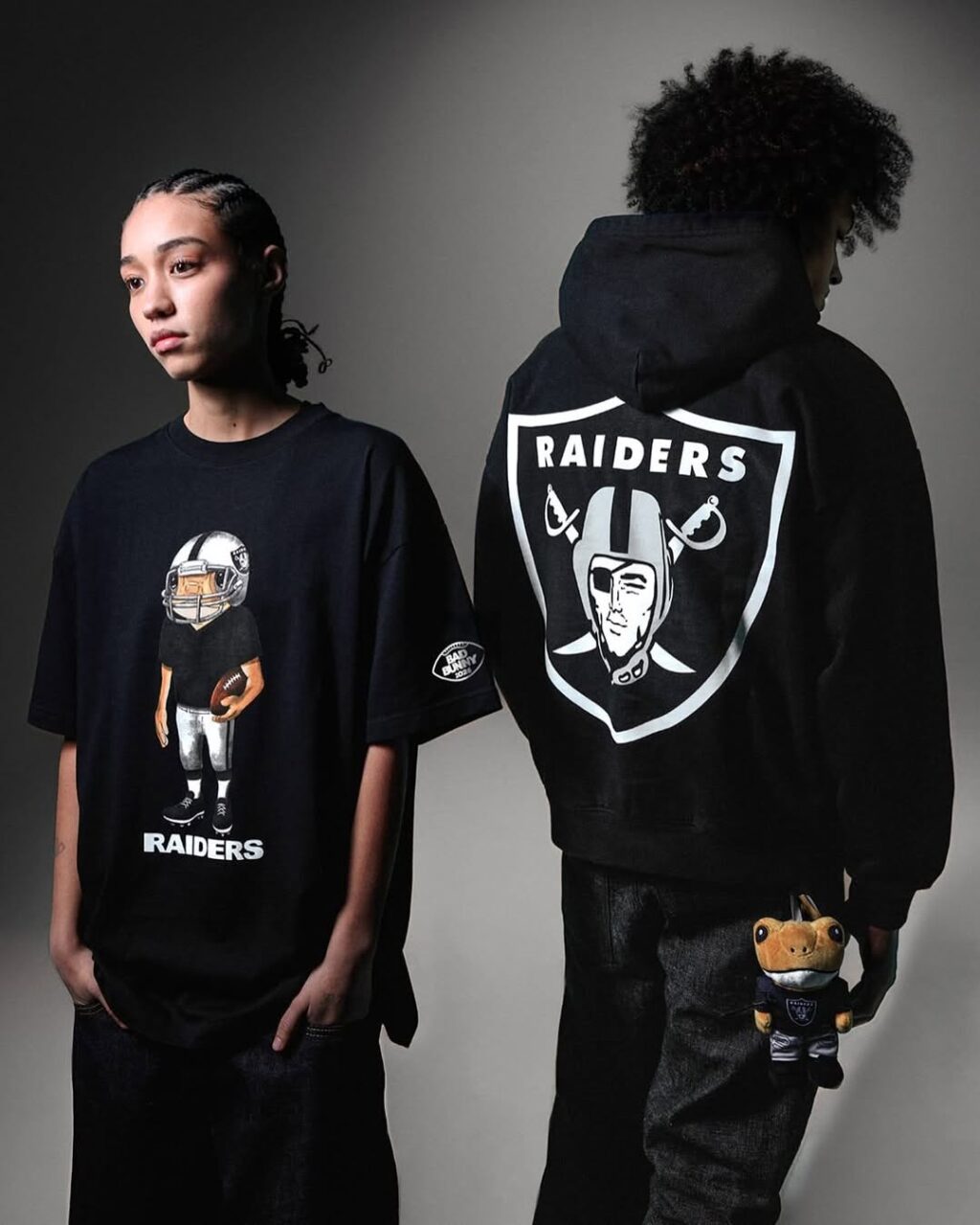 Dois jovens usando roupas da coleção NFL e Bad Bunny, um com camiseta preta ilustrada e outro com moletom e chaveiro. Ambos exibem o logotipo do time RAIDERS.