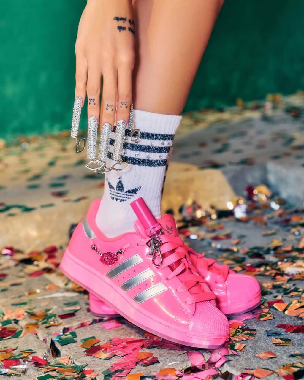 Detalhe de um par de tênis Adidas Superstar rosa com detalhes prateados, exibidos ao lado de uma mão com unhas longas, decoradas com strass e anéis, em um ambiente com confetes coloridos pelo chão.