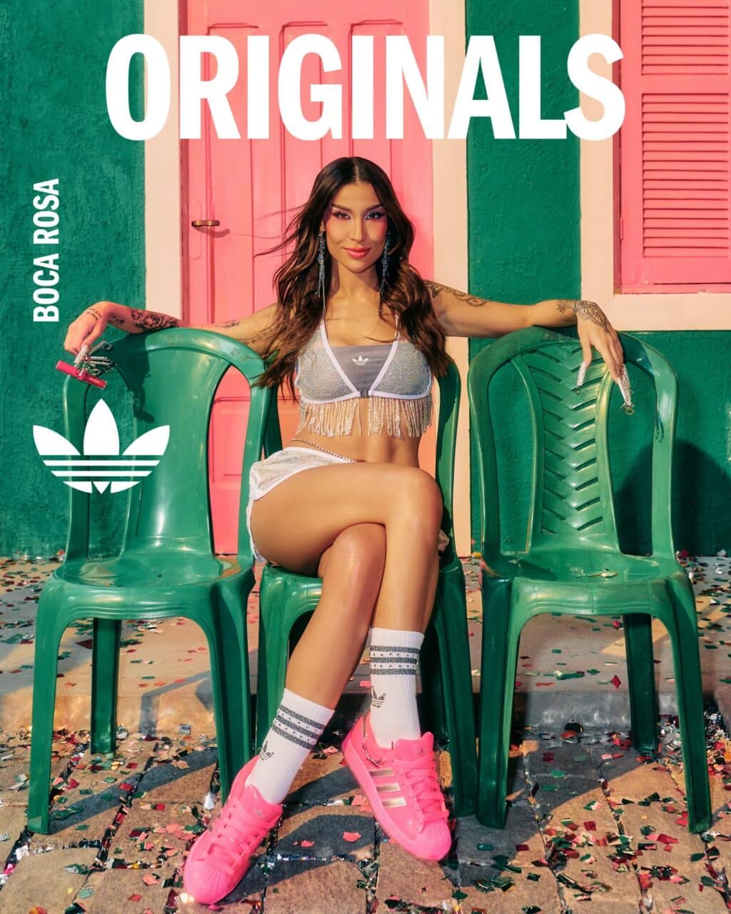 Modelo em trajes da coleção Originals da collab entre Adidas e Boca Rosa, sentada em cadeiras plásticas verdes, com decoração de carnaval ao fundo.