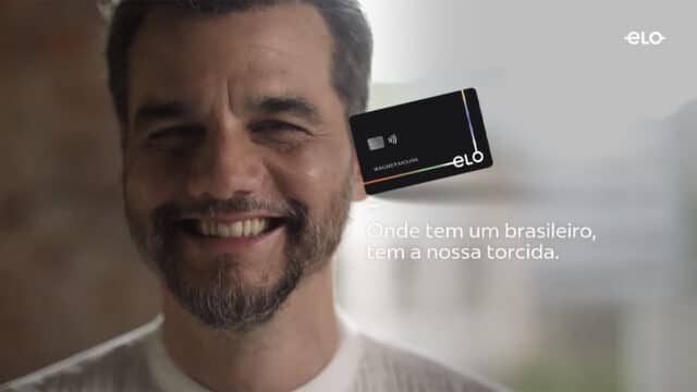 Ator Wagner Moura sorrindo ao lado de um cartão Elo e a frase "Onde tem um brasileiro, tem a nossa torcida"