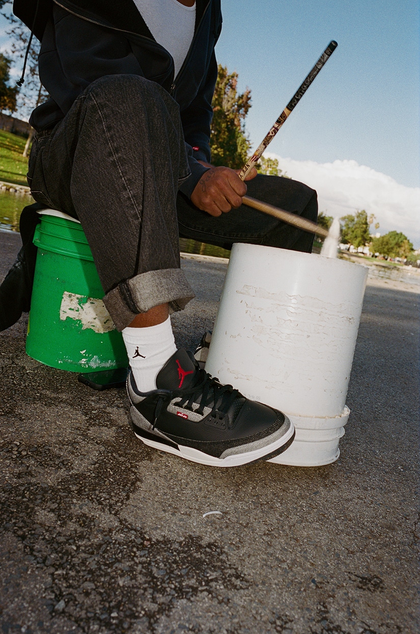 Close-up de uma pessoa tocando bateria em um balde branco enquanto usa tênis Air Jordan 3 pretos e vermelhos da colaboração Levi's x Jordan Brand.