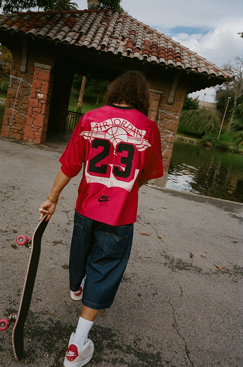 Pessoa caminhando em um ambiente ao ar livre, usando uma camisa vermelha da Air Jordan com o número 23 nas costas, calças jeans largas e segurando um skate.