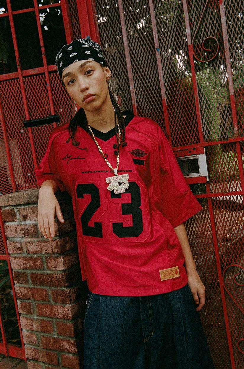 Uma modelo usando uma camisa de futebol americano vermelha com o número 23, acessórios de corrente e um bandana, posando em frente a uma grade vermelha.
