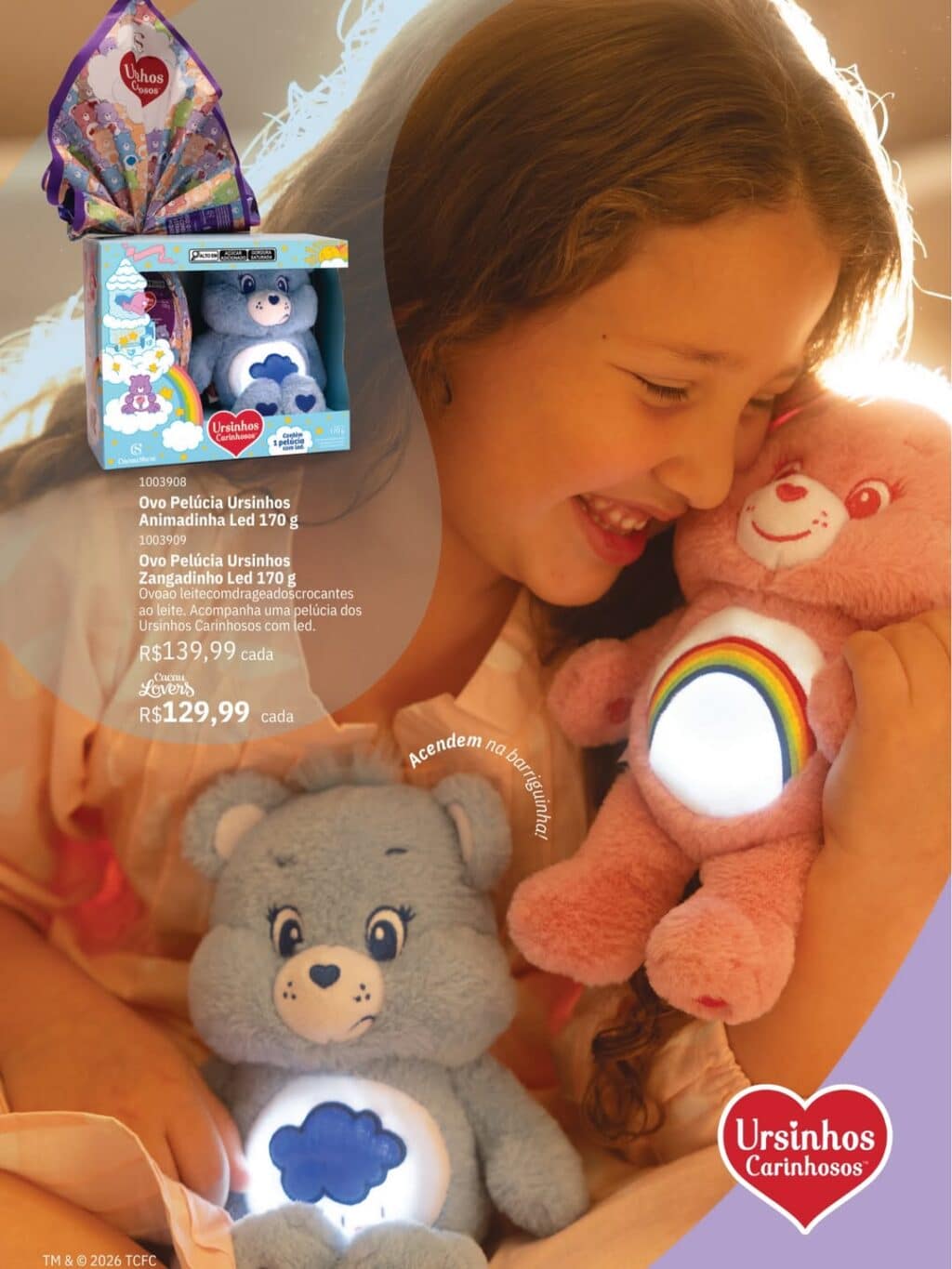 Uma criança sorri enquanto segura os Ursinhos Carinhosos, um azul chamado Zangadinho com barriguinha iluminada, e um rosa chamado Animadinha. Ao fundo, uma caixa decorativa apresenta os produtos da Cacau Show para a Páscoa 2026.