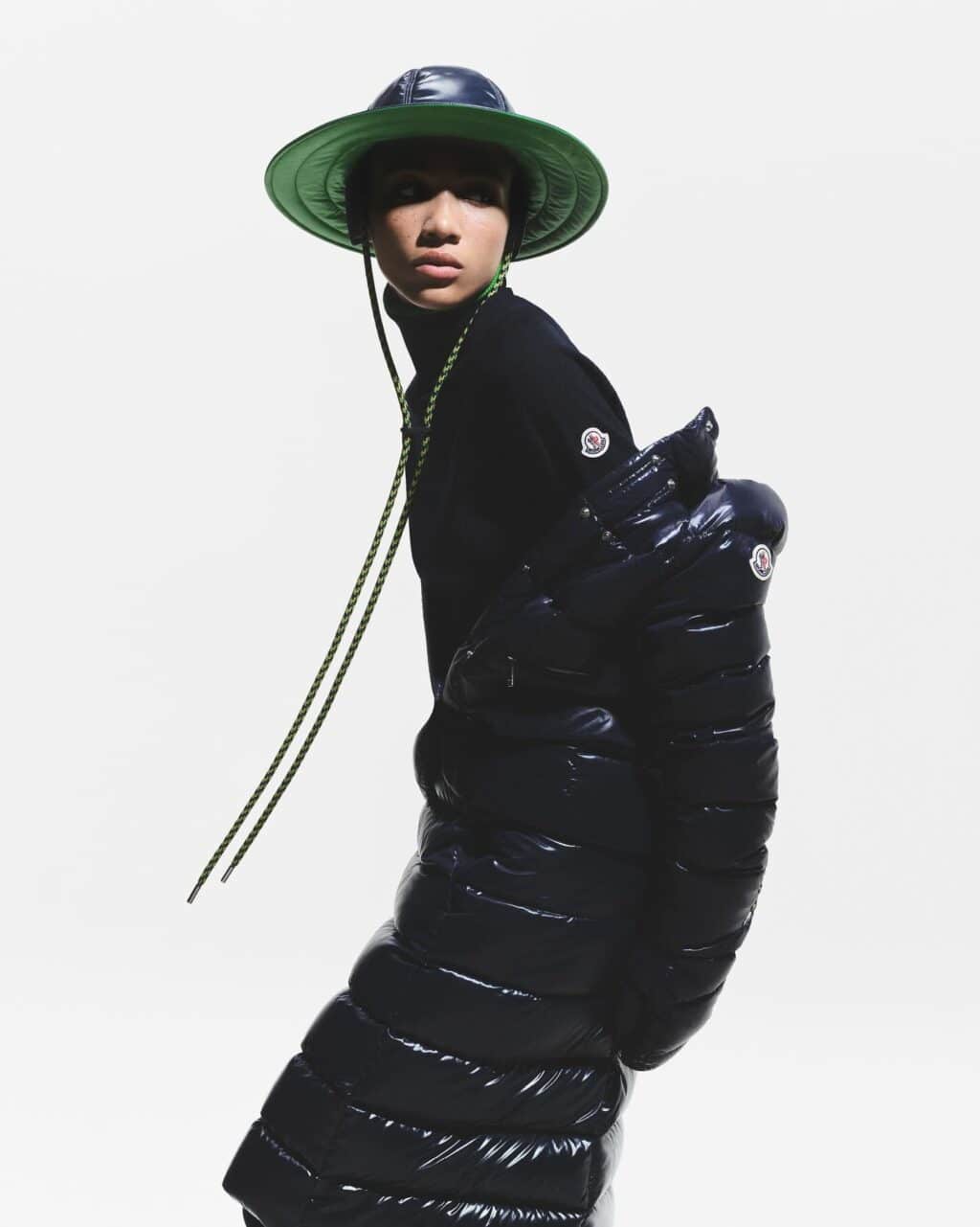 Uma modelo vestindo um casaco puffer preto brilhante e um chapéu verde de aba larga, posou contra um fundo branco.