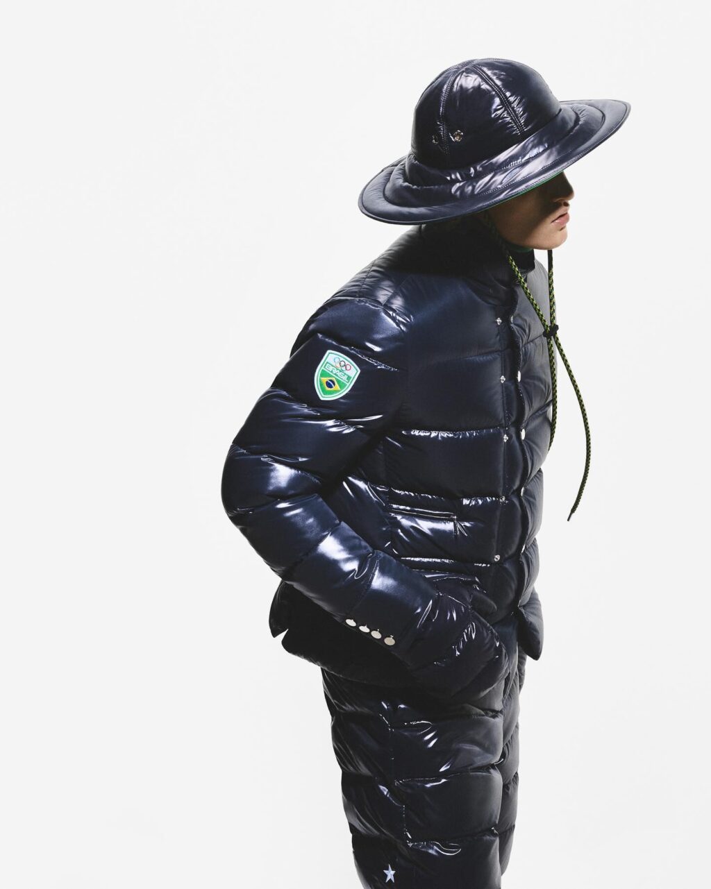 Uma modelo vestindo uma jaqueta puffer azul-marinho brilhante com um distintivo e um chapéu de aba larga, em pé contra um fundo branco.