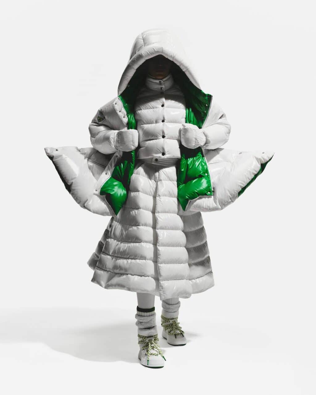 Uma modelo vestindo um elegante casaco puffer oversized com exterior branco brilhante e interior verde vibrante, com capuz e fechamento por botões, combinado com calças puffer da mesma cor e tênis robustos.