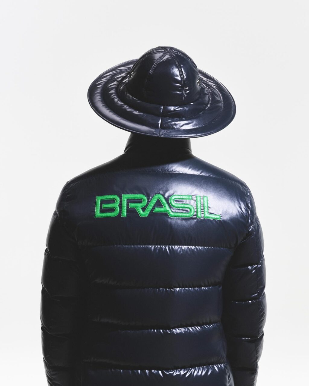 Uma pessoa vestindo uma jaqueta puffer escura e brilhante com a palavra 'BRASIL' bordada em verde nas costas e um chapéu de aba larga, vista de costas contra um fundo branco.
