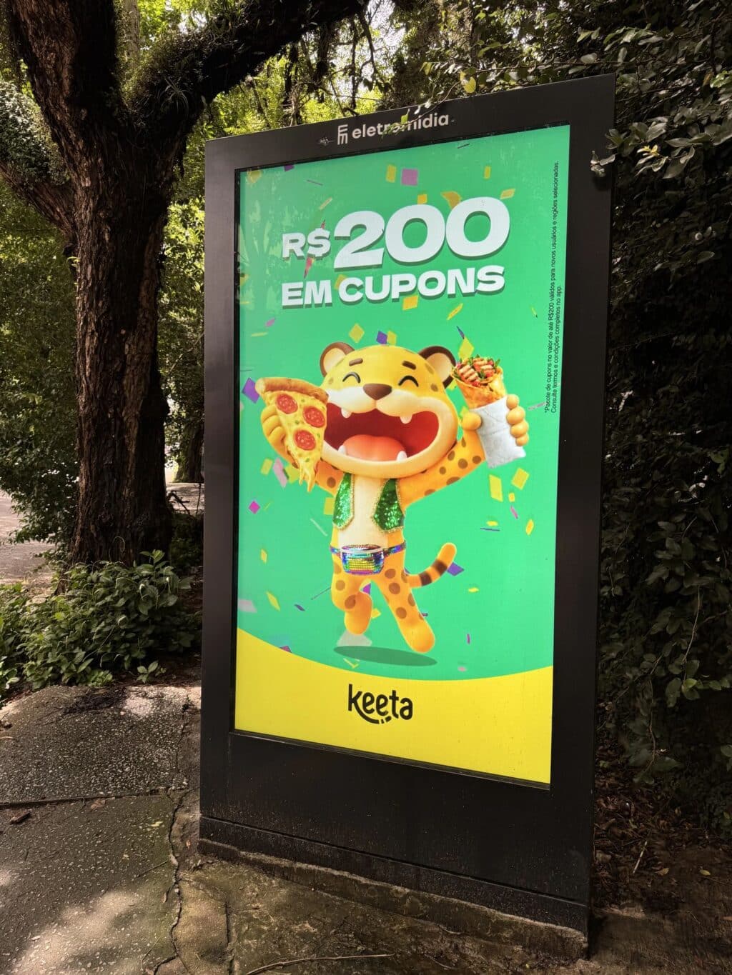 Um anúncio colorido com um tigre de desenho animado segurando pizza e sorvete, promovendo cupons de desconto de R$ 200 para a Keeta. O fundo é verde vibrante com confetes.