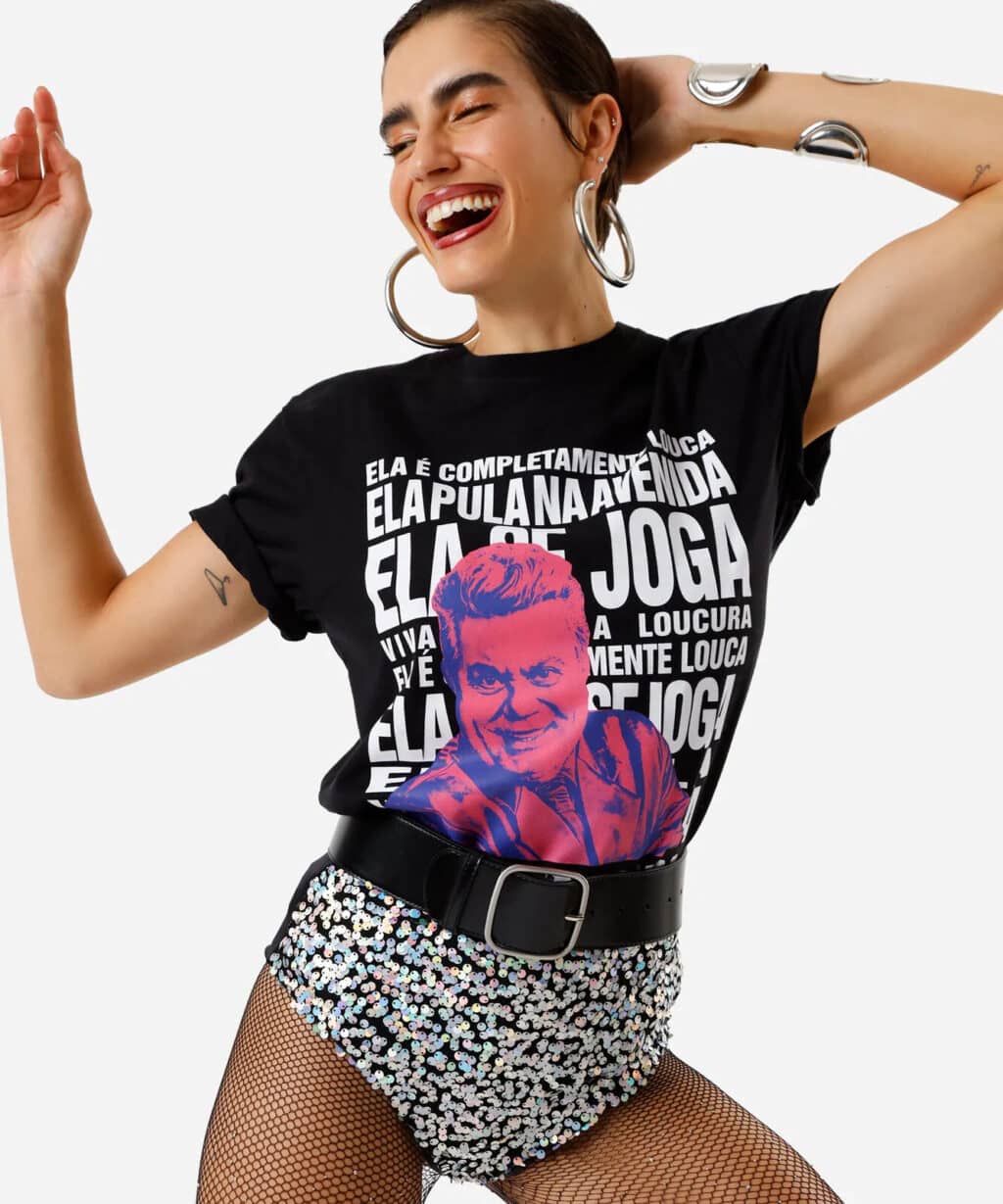 Modelo sorridente vestindo uma camiseta preta com frases e uma ilustração colorida, acessórios grandes e saia brilhante, em pose animada.