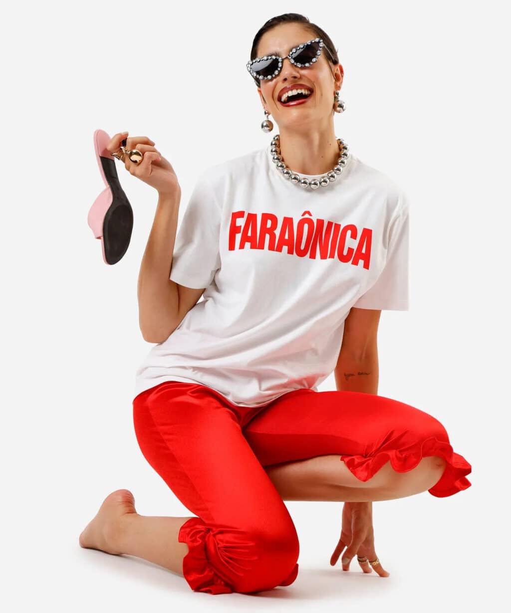 Modelo feminina sorrindo, vestindo uma camiseta branca com a palavra 'FARAÔNICA' em destaque vermelho, calças vermelhas com detalhes e acessórios glamourosos, incluindo óculos escuros e um colar de pérolas, enquanto segura um sapato de salto rosa.