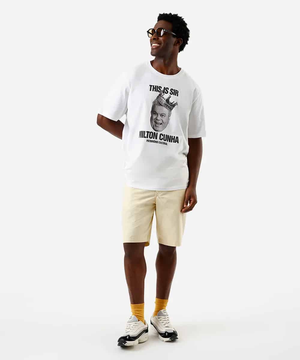 Um homem sorridente usando uma camiseta branca com a frase 'THIS IS SIR MILTON CUNHA' e uma imagem do comunicador, combinado com shorts claros e tênis, em um fundo neutro.