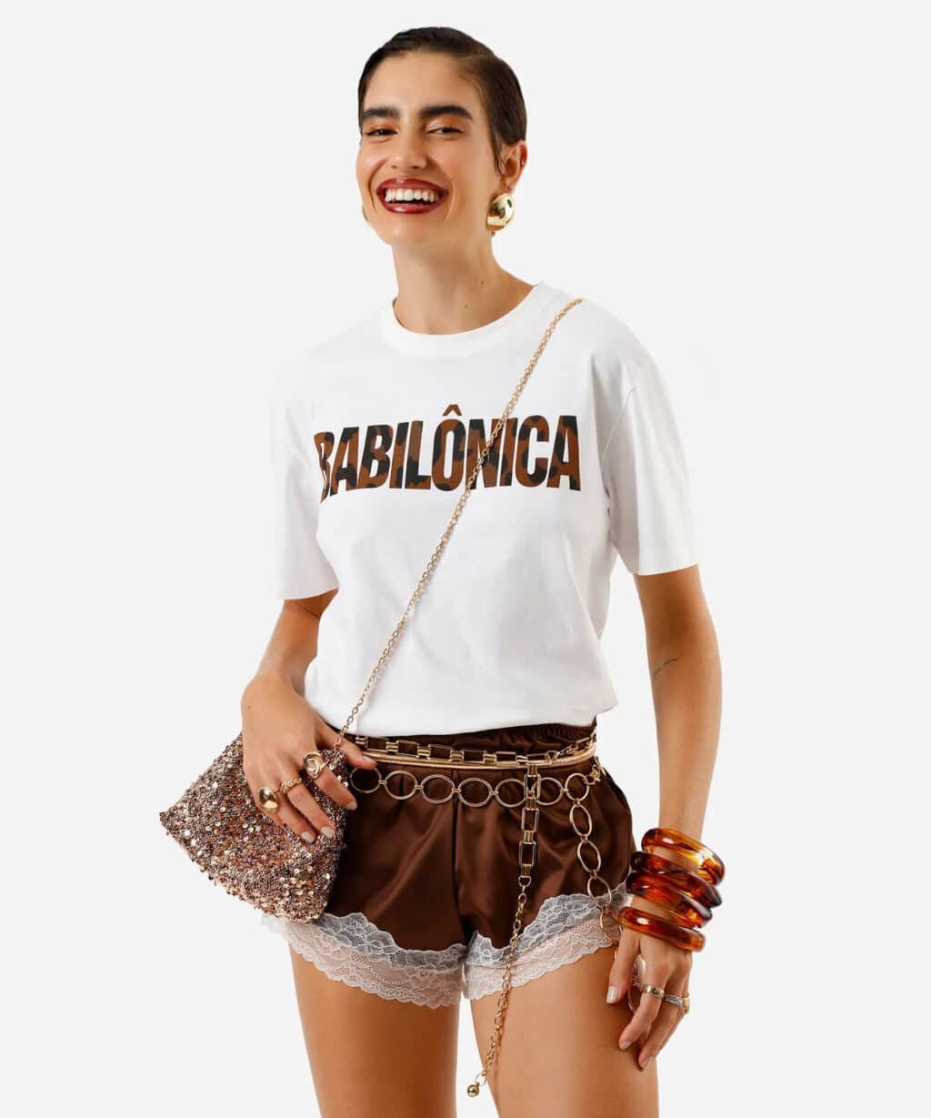 Modelo sorridente usando uma camiseta branca com a palavra 'BABILÔNICA' estampada em letras grandes e escuras, acompanhada de shorts marrons com detalhes em renda. Acessórios incluem anéis, pulseiras de acrílico, e uma bolsa de mão brilhante.