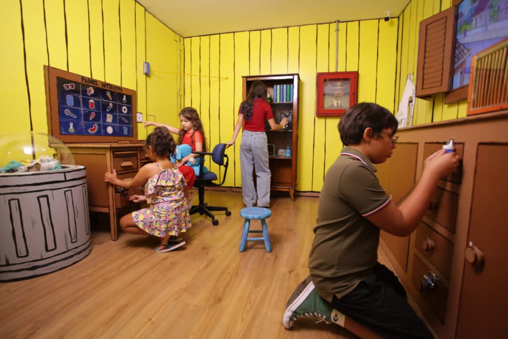 Uma sala de jogos colorida com quatro crianças interagindo com os móveis e um quadro-negro, em um cenário de paredes amarelas vibrantes.