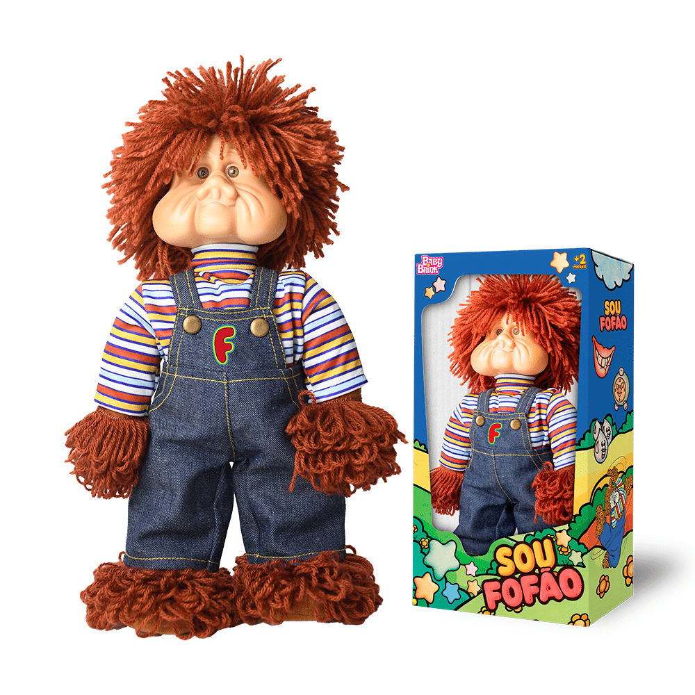Uma boneca peculiar com uma cabeça grande, cabelo ruivo encaracolado e um rosto sorridente, vestindo uma camisa listrada e um macacão jeans, exposta ao lado de sua embalagem colorida.