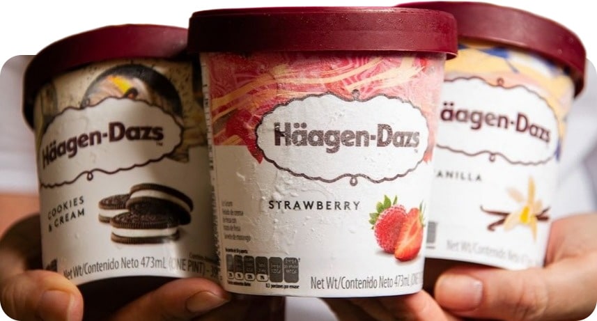 Três potes de sorvete Häagen-Dazs nos sabores Cookies & Cream, Morango e Baunilha, nas mãos de uma pessoa.