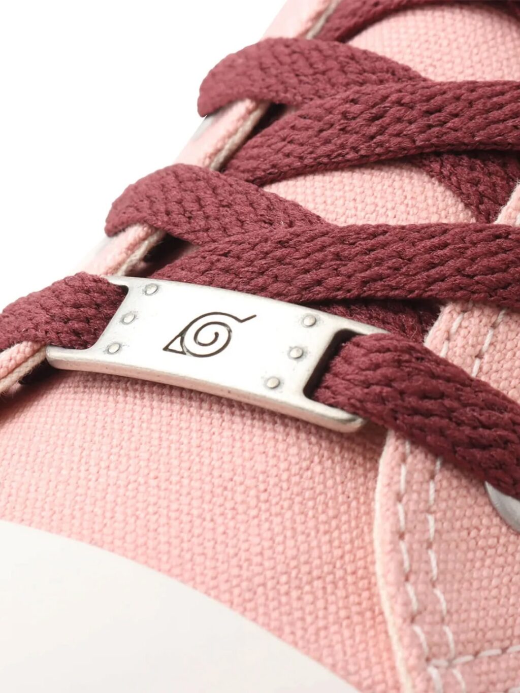 Detalhe de um tênis rosa com cadarços cor de vinho, apresentando uma etiqueta metálica com um símbolo circular.