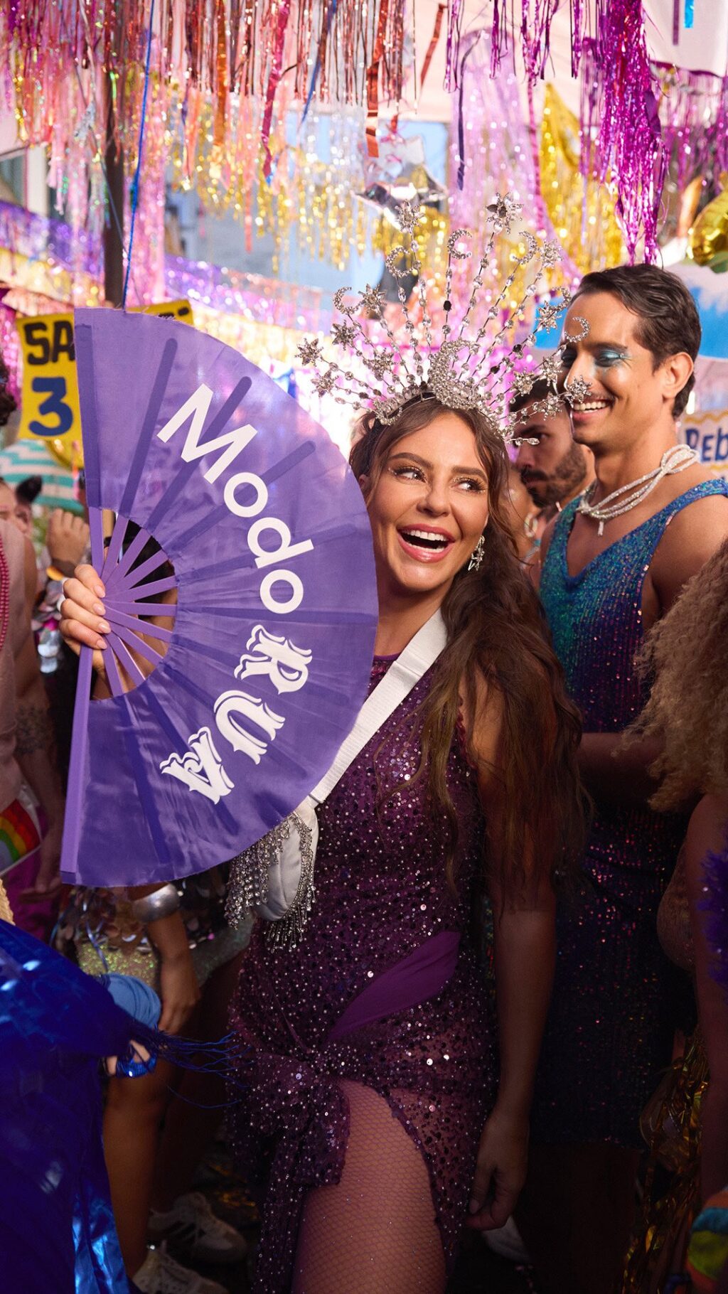 A joyful woman in a sparkling purple dress and adorned with a crown holds a purple fan that says 'Modo Rua,' celebrating in a vibrant crowd during a festive event.