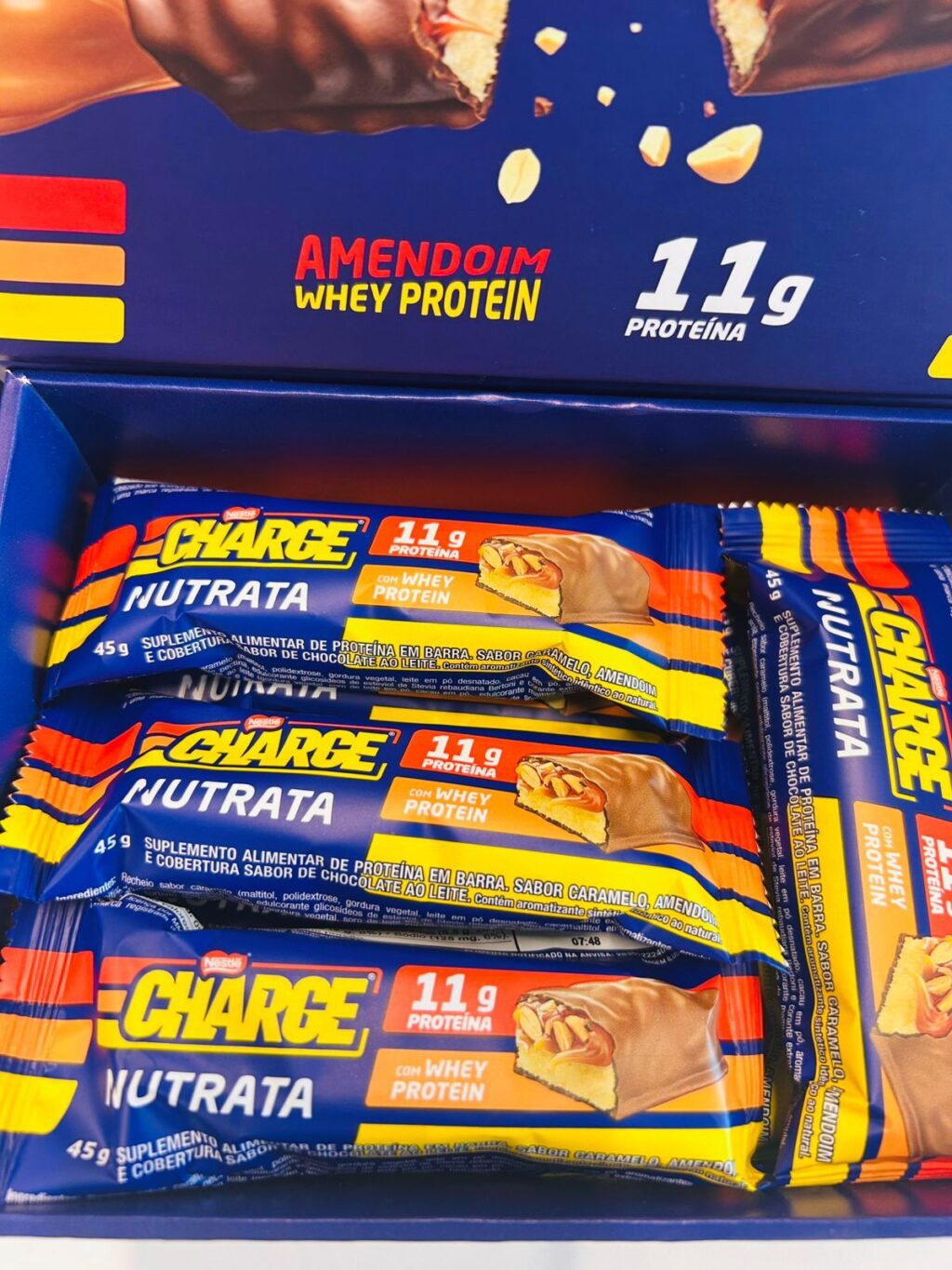 Uma caixa de barras de proteína com diversos sabores, destacando as informações "11 g de proteína" e "proteína do soro do leite". O sabor em destaque é amendoim, com imagens das barras e seus ingredientes.
