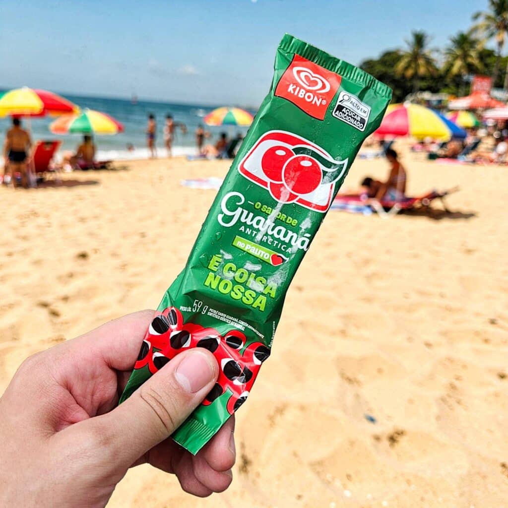 Picolé de guaraná Kibon segurado por uma mão, com a praia ao fundo e pessoas relaxando sob guarda-sóis coloridos.