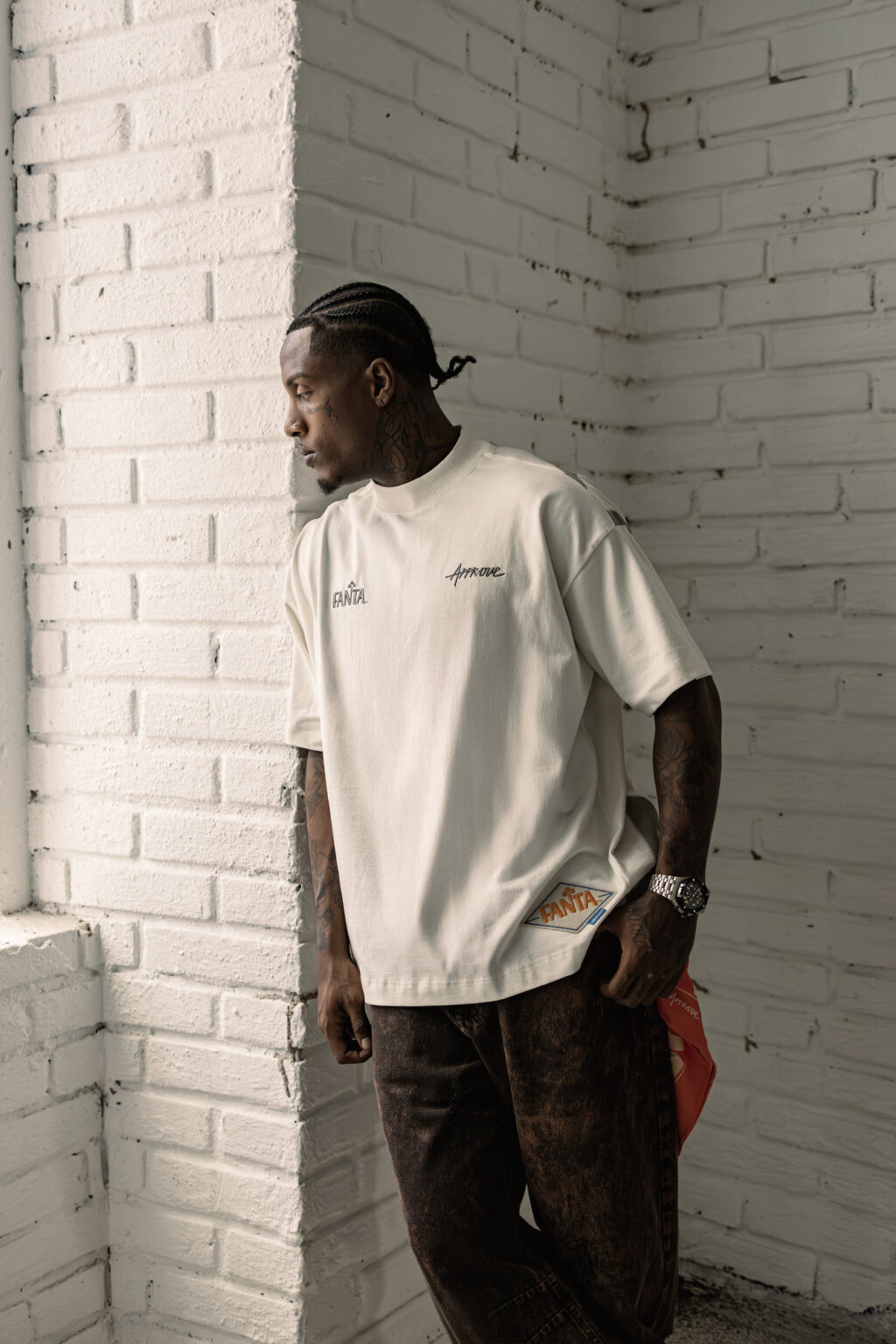 A person leaning against a white brick wall, wearing a light-colored oversized t-shirt with 'Fanta' branding and the word 'Approve' embroidered on it, paired with dark, patterned pants.