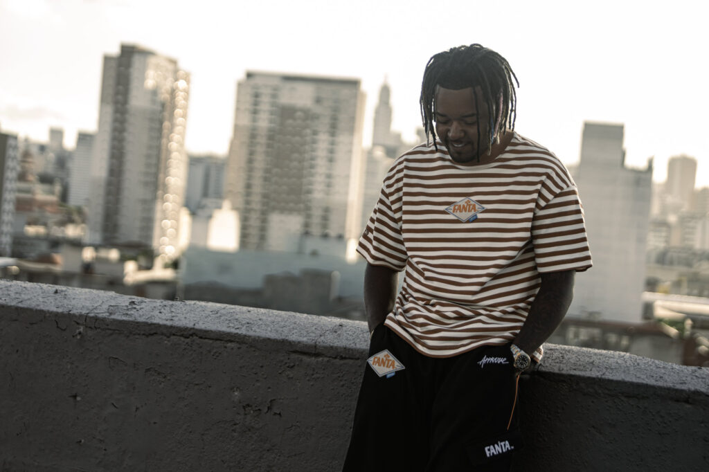 A man with dreadlocks wearing a brown and white striped t-shirt and black shorts stands on a rooftop, looking down with a city skyline in the background.